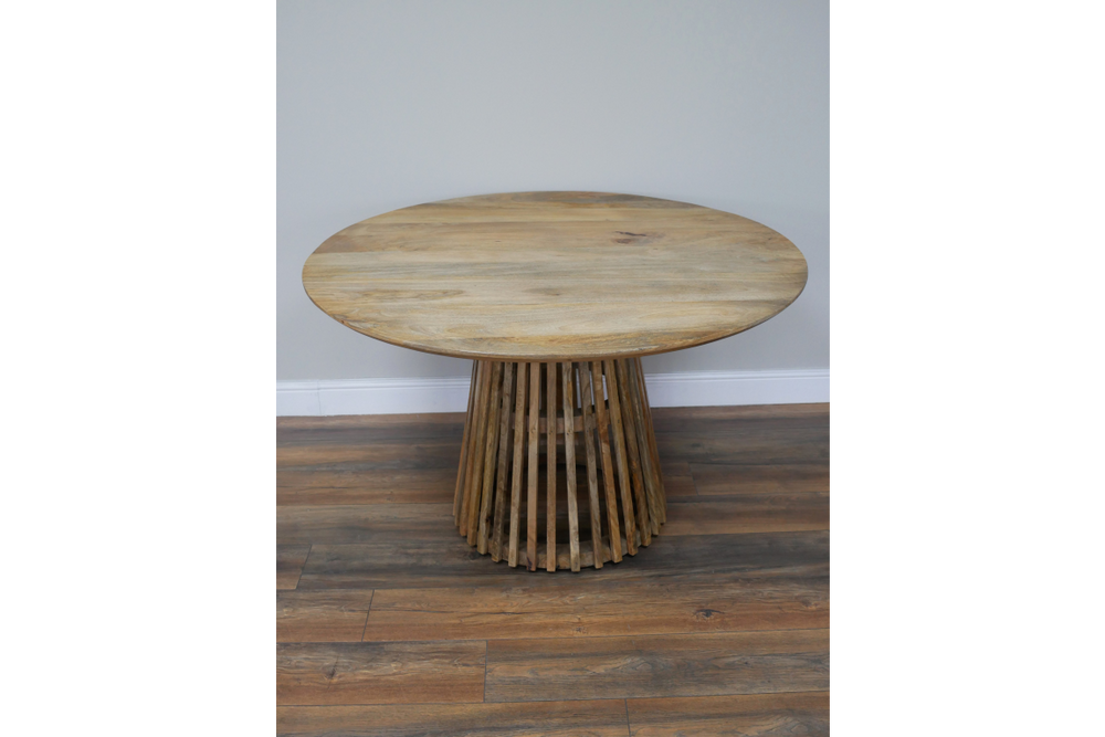 Round wooden dining table with slatted base, natural finish, front view on wooden floor against gray wall.