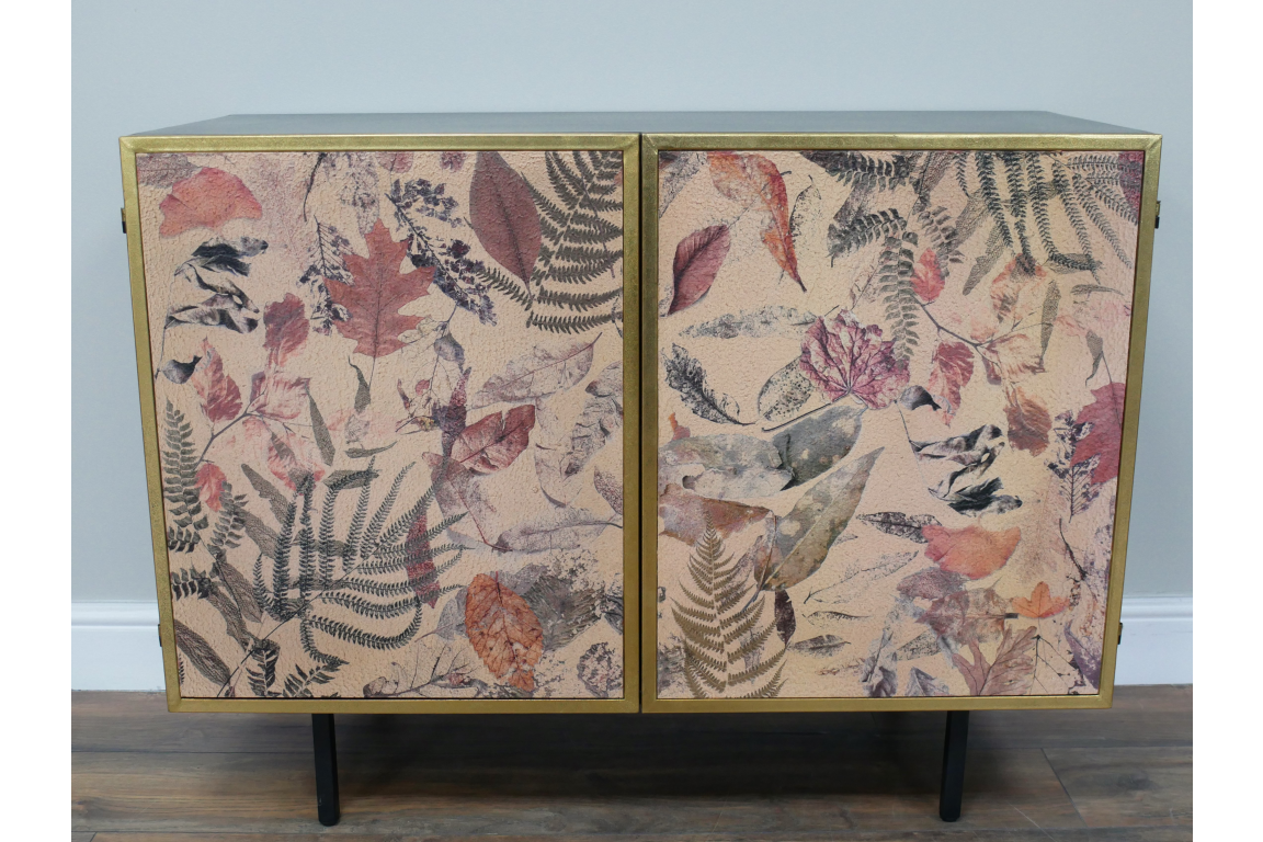 Small cabinet with two doors featuring autumn leaf and fern print, gold frame, black legs, against gray wall on wood floor.