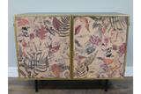 Small cabinet with two doors featuring autumn leaf and fern print, gold frame, black legs, against gray wall on wood floor.