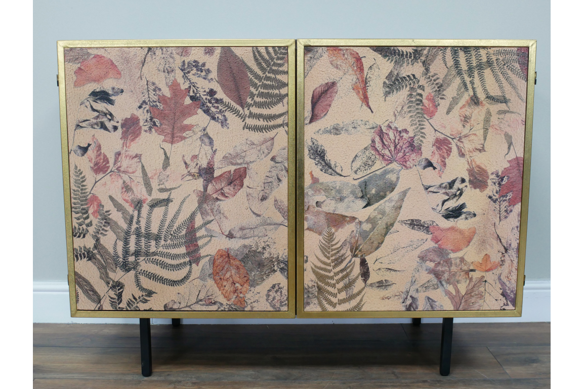 Small cabinet with two doors, black metal legs, gold frame, and leaf-patterned front panels in earthy tones.