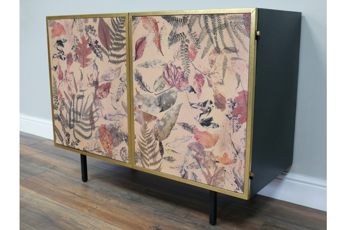Small black metal cabinet with gold-framed doors featuring pink and purple pressed leaf and fern print, angled view.