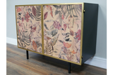 Small black metal cabinet with gold-framed doors featuring pink and purple pressed leaf and fern print, angled view.