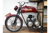 Metal and wood motorbike-shaped bar cabinet in red and black, side view, indoors on wooden floor near shelving unit.