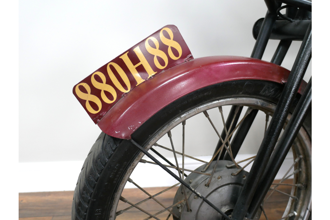 Close-up angle of motorbike bar cabinet wheel with black metal spokes, maroon fender, and yellow lettering plate.