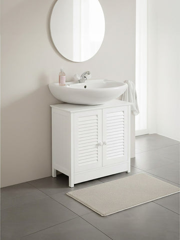 White wood under sink bathroom cabinet with shutter-style doors, featuring a round mirror above and a modern basin sink. A soft beige rug is placed on the floor in front of the cabinet, set against a light-colored wall and tiled flooring