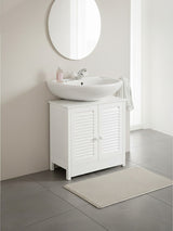 White wood under sink bathroom cabinet with shutter-style doors, featuring a round mirror above and a modern basin sink. A soft beige rug is placed on the floor in front of the cabinet, set against a light-colored wall and tiled flooring