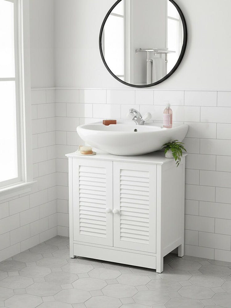 White wood under sink bathroom cabinet with shutter-style doors, a round mirror above, and a modern sink on top. The cabinet is positioned against a light-colored wall with a window, and a small plant and toiletries are placed on the sink. The floor is tiled in a light gray color