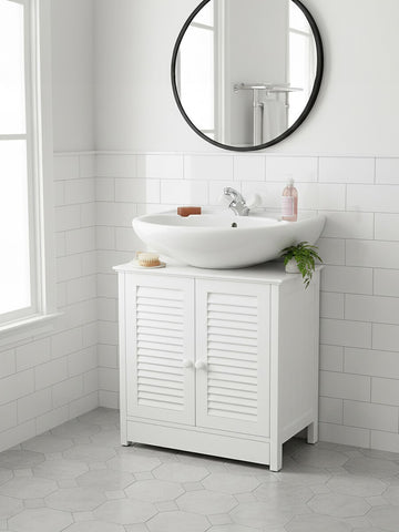 White wood under sink bathroom cabinet with shutter-style doors, a round mirror above, and a modern sink on top. The cabinet is positioned against a light-colored wall with a window, and a small plant and toiletries are placed on the sink. The floor is tiled in a light gray color