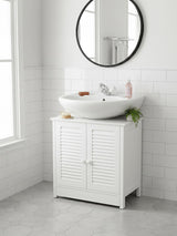 White wood under sink bathroom cabinet with shutter-style doors, a round mirror above, and a modern sink on top. The cabinet is positioned against a light-colored wall with a window, and a small plant and toiletries are placed on the sink. The floor is tiled in a light gray color