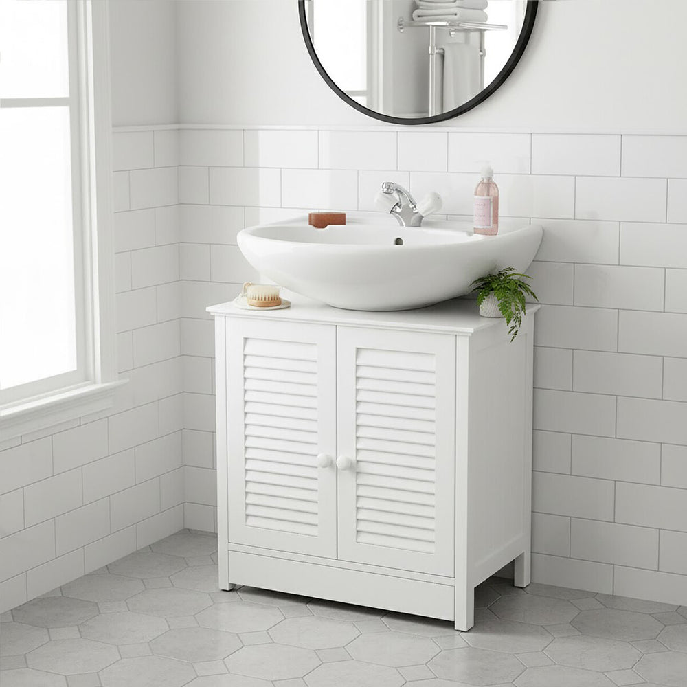 A white wood under sink bathroom cabinet with shutter-style doors, featuring a round mirror above it. A modern, circular sink sits on top of the cabinet, accompanied by a faucet and decorative items. The cabinet is positioned against a tiled wall and has a clean, minimalist design. Natural light streams in from a nearby window, illuminating the space