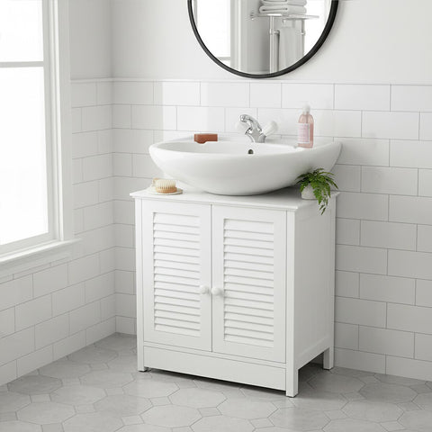 A white wood under sink bathroom cabinet with shutter-style doors, featuring a round mirror above it. A modern, circular sink sits on top of the cabinet, accompanied by a faucet and decorative items. The cabinet is positioned against a tiled wall and has a clean, minimalist design. Natural light streams in from a nearby window, illuminating the space