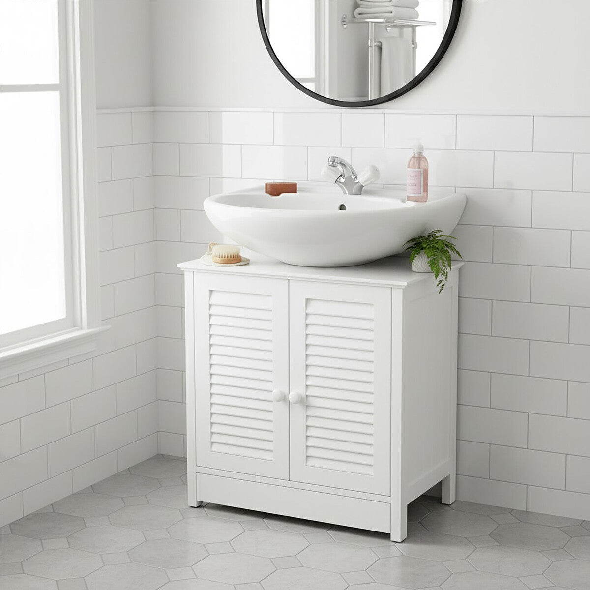 A white wood under sink bathroom cabinet with shutter-style doors, featuring a round mirror above it. A modern, circular sink sits on top of the cabinet, accompanied by a faucet and decorative items. The cabinet is positioned against a tiled wall and has a clean, minimalist design. Natural light streams in from a nearby window, illuminating the space