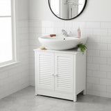 A white wood under sink bathroom cabinet with shutter-style doors, featuring a round mirror above it. A modern, circular sink sits on top of the cabinet, accompanied by a faucet and decorative items. The cabinet is positioned against a tiled wall and has a clean, minimalist design. Natural light streams in from a nearby window, illuminating the space