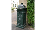 Green cast iron post box with ornate detailing beside brick wall and garden hedge