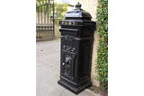 Black cast iron post box with ornate detailing beside stone wall and garden gate