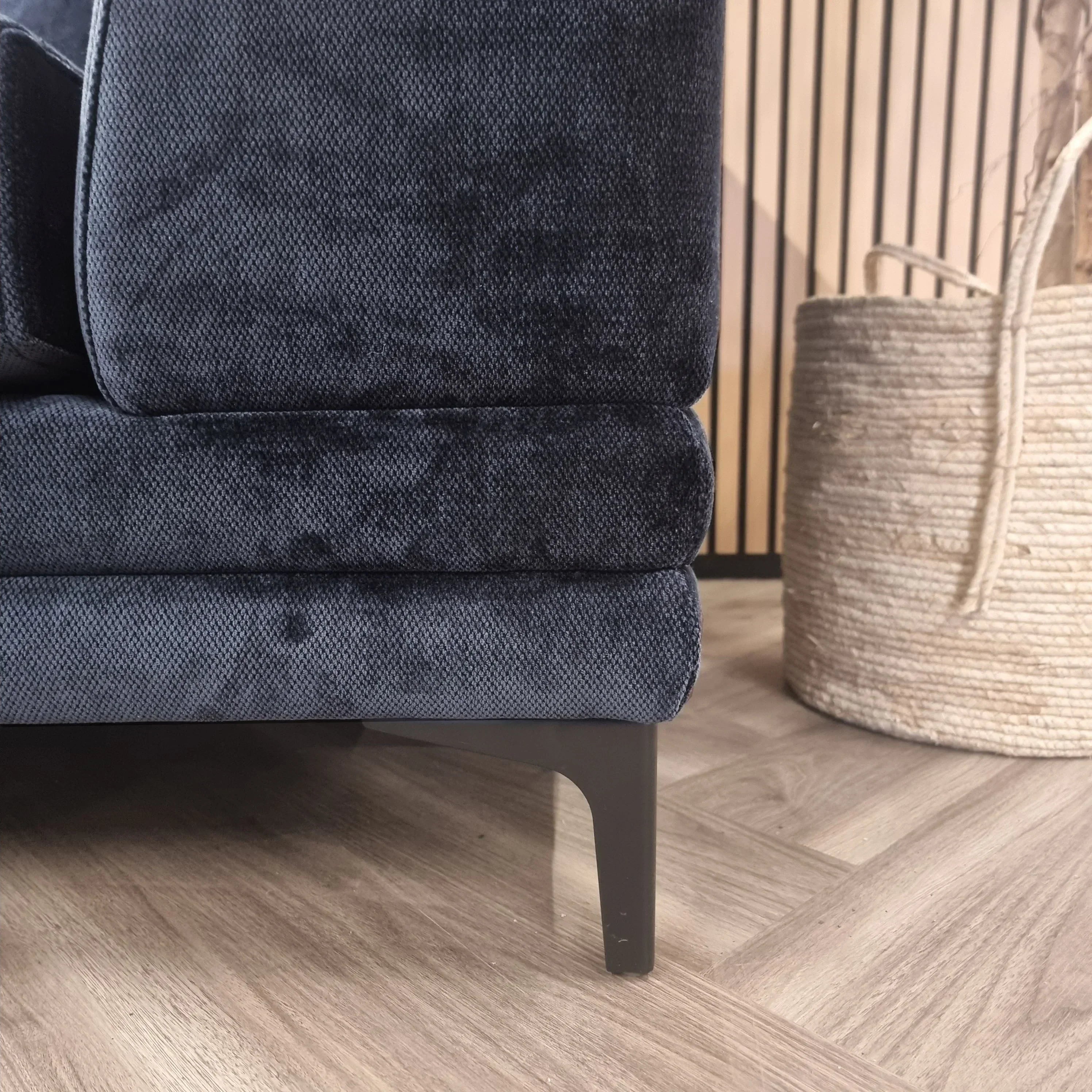 Dark fabric sofa corner with black metal leg on wood floor, basket planter in background