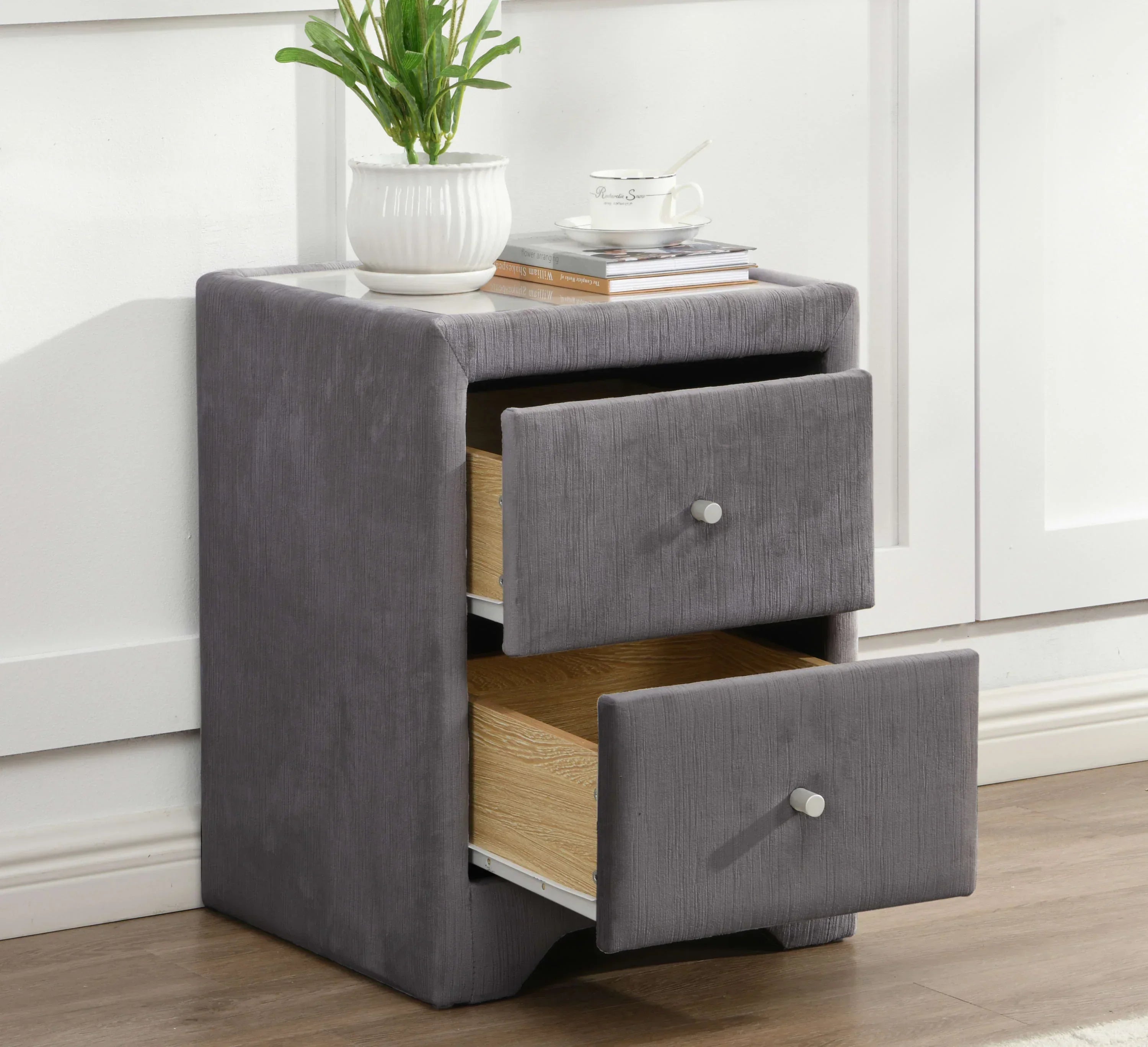 Grey fabric bedside table with two open drawers, potted plant and cup on top