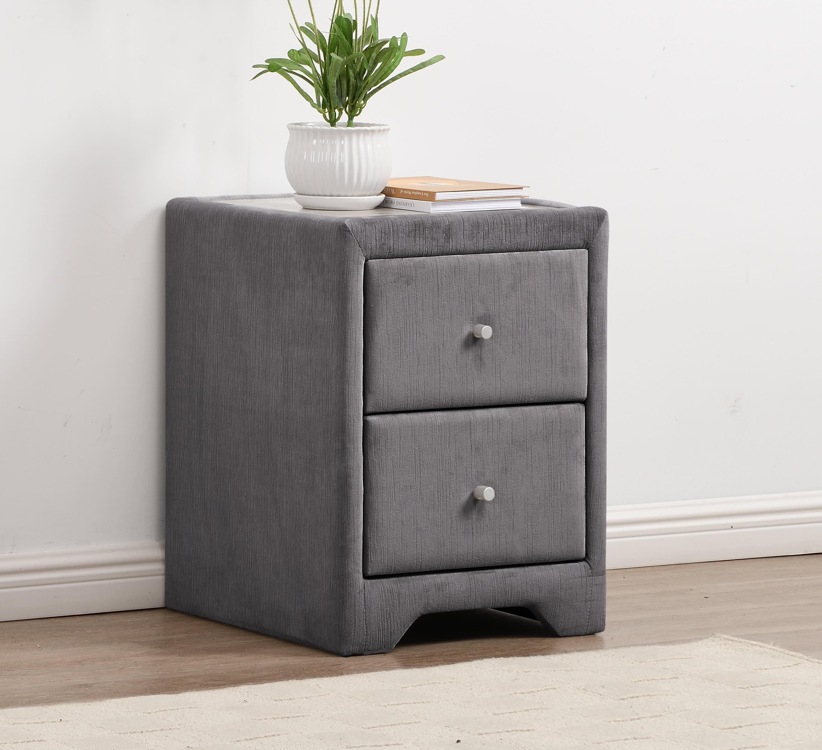 Grey fabric bedside table with two drawers and a plant on top, set against a white wall