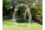 Metal garden arch with ornate scrollwork on grass lawn surrounded by plants and trees