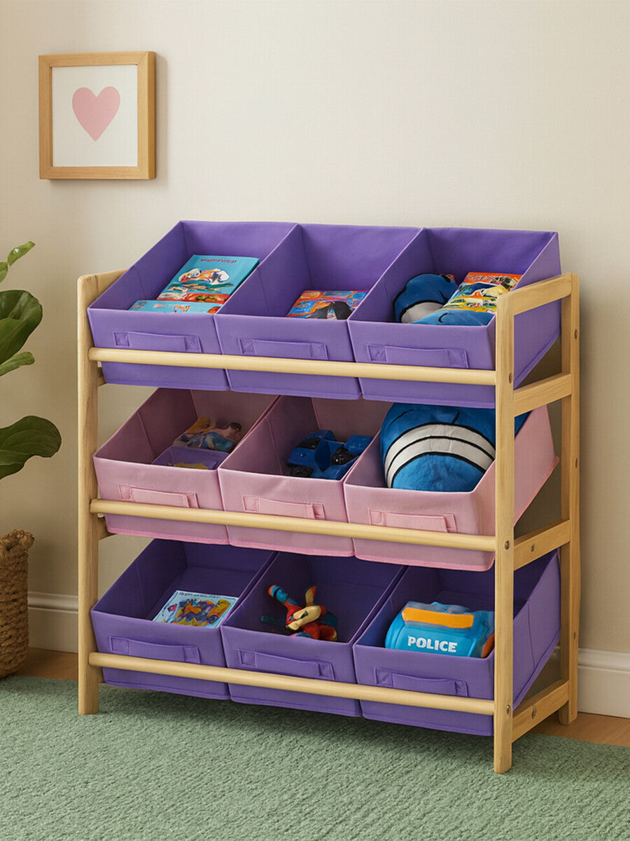A three-tier kids storage unit featuring a wooden frame with light natural wood finish. The top two tiers have purple fabric bins, while the bottom tier has a pink fabric bin. Each bin is filled with colorful books and toys, and the unit is placed against a light-colored wall with a framed heart artwork above it. The floor is covered with a green rug