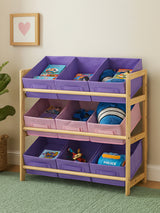 A three-tier kids storage unit featuring a wooden frame with light natural wood finish. The top two tiers have purple fabric bins, while the bottom tier has a pink fabric bin. Each bin is filled with colorful books and toys, and the unit is placed against a light-colored wall with a framed heart artwork above it. The floor is covered with a green rug