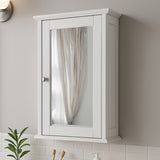 A white mirrored cabinet mounted on a wall, featuring a single door with a mirror front and a small handle. The cabinet has a simple, modern design and is positioned above a countertop with a plant and other decorative items visible below. Soft lighting illuminates the scene, creating a warm ambiance
