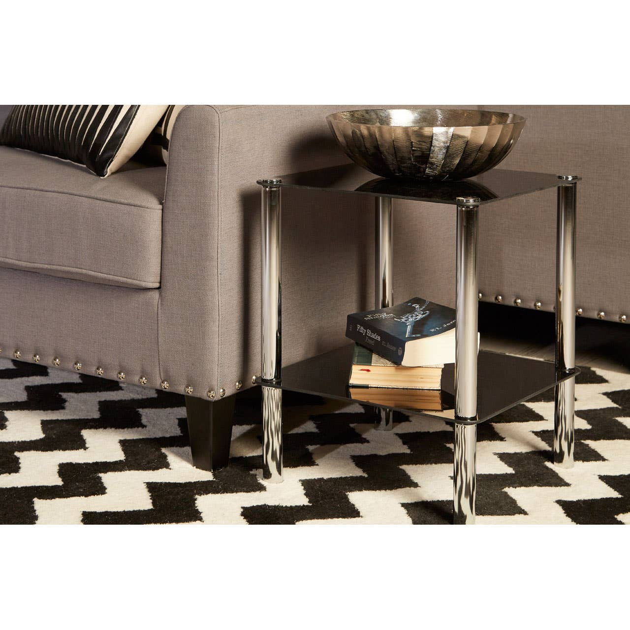 A black glass end table with two tiers, featuring sleek metal legs, positioned next to a gray sofa. The table holds a decorative bowl and a stack of books, set on a black and white zigzag patterned rug