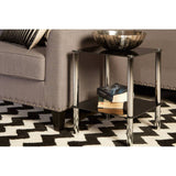 A black glass end table with two tiers, featuring sleek metal legs, positioned next to a gray sofa. The table holds a decorative bowl and a stack of books, set on a black and white zigzag patterned rug