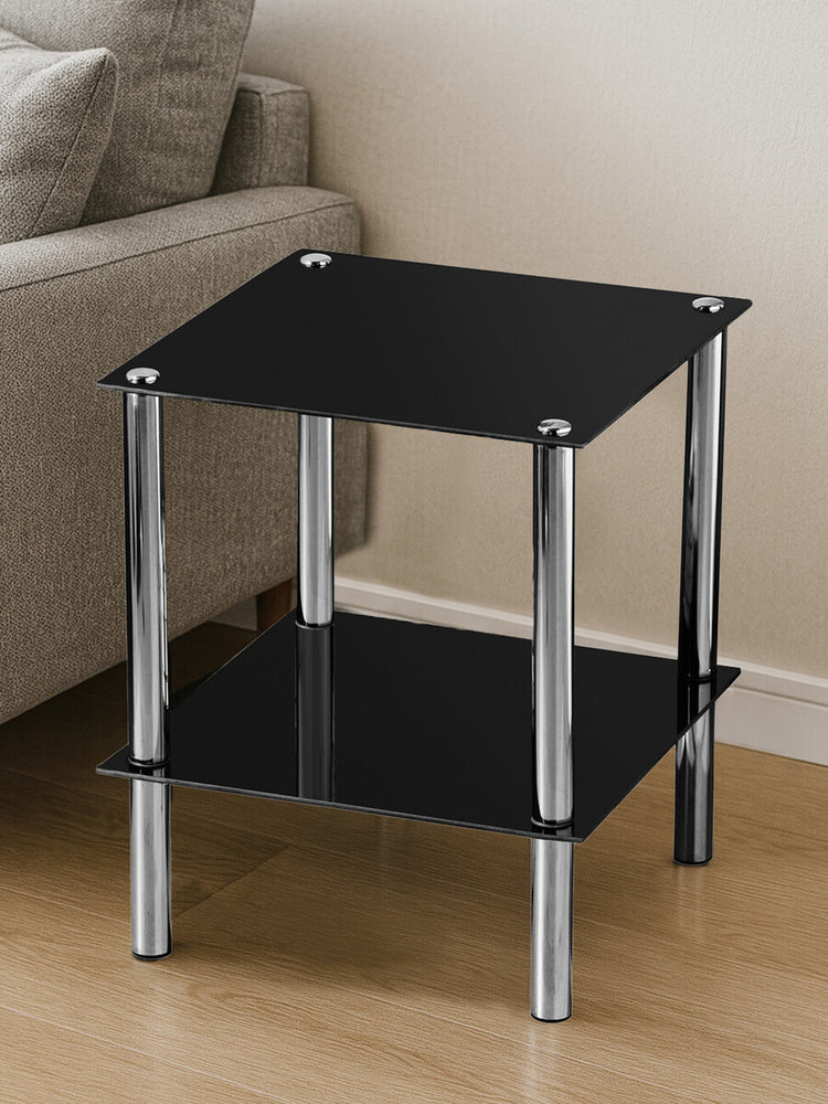 Black glass end table with two tiers, featuring a sleek design and chrome legs, placed on a light wooden floor next to a beige sofa