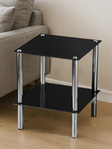 Black glass end table with two tiers, featuring a sleek design and chrome legs, placed on a light wooden floor next to a beige sofa