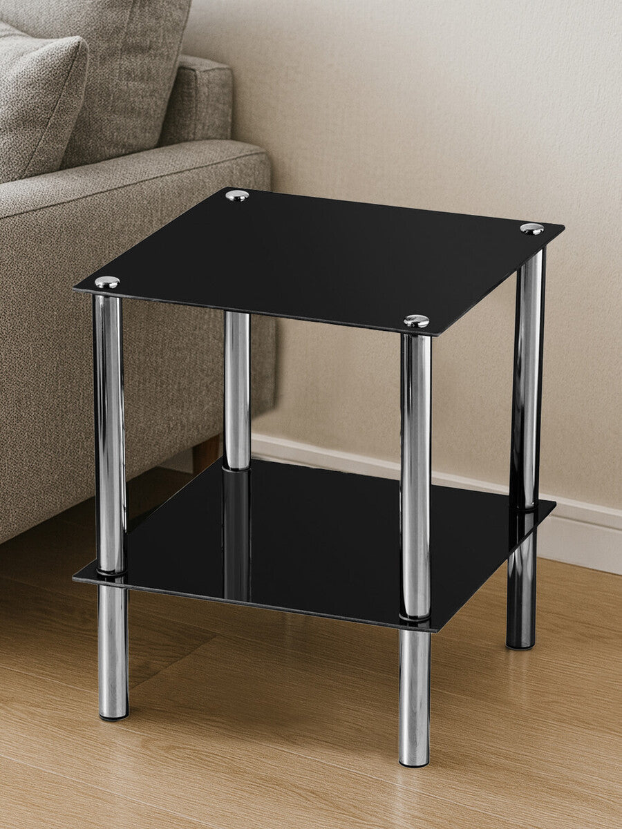 Black glass end table with two tiers, featuring a sleek design and chrome legs, placed on a light wooden floor next to a beige sofa