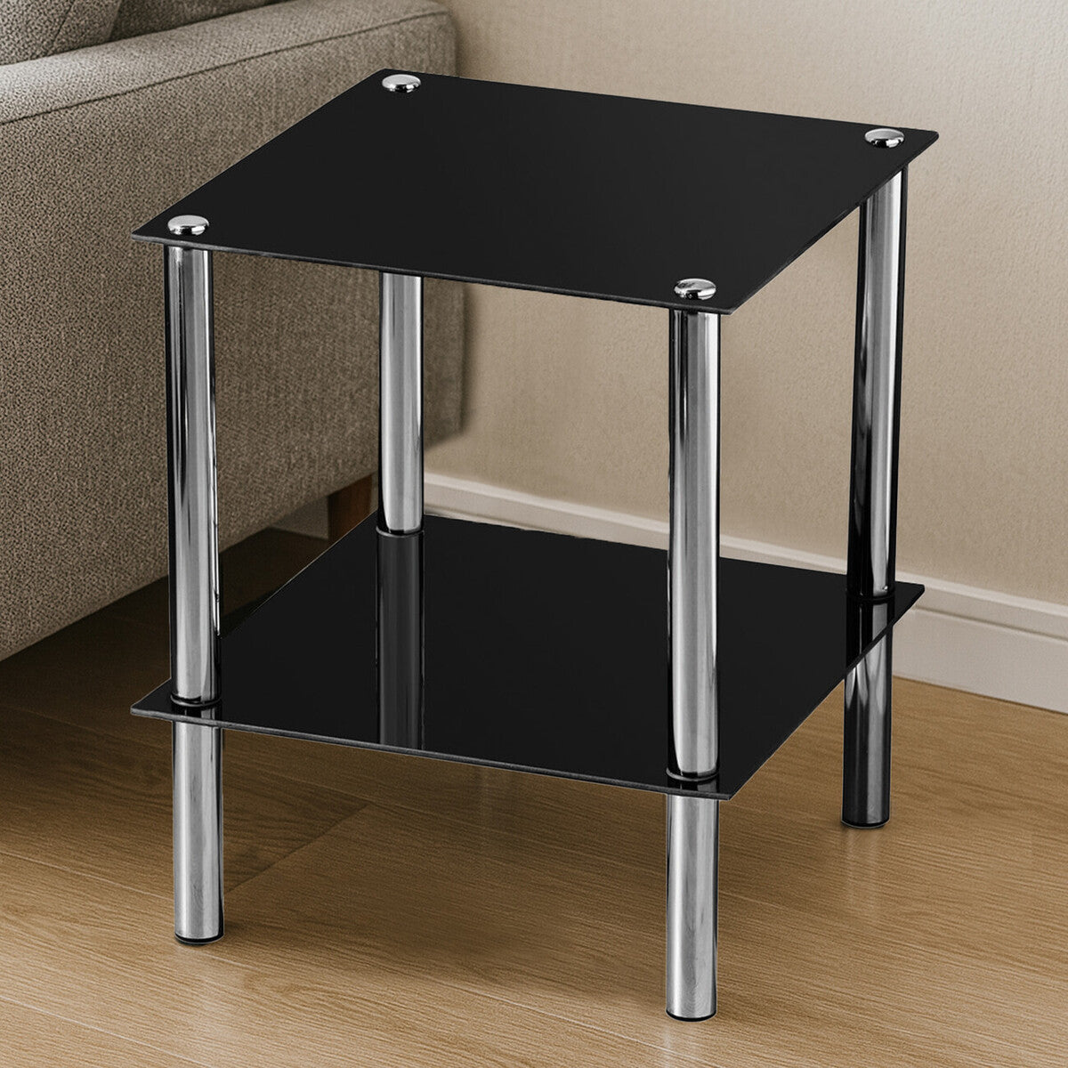 Black glass end table with two tiers, featuring a sleek design and chrome legs, placed on a light wooden floor next to a sofa