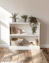 A white wooden storage unit with two open shelves, displaying a few books, a decorative bowl, and several potted plants. Sunlight casts soft shadows on the floor, highlighting the natural wood grain. The background features a light-colored wall and wooden flooring