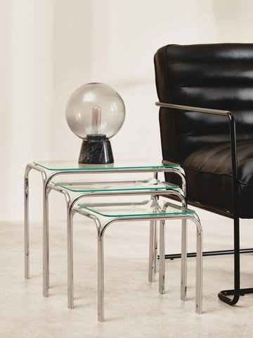 Glass nesting tables with chrome legs beside black leather armchair in modern living room