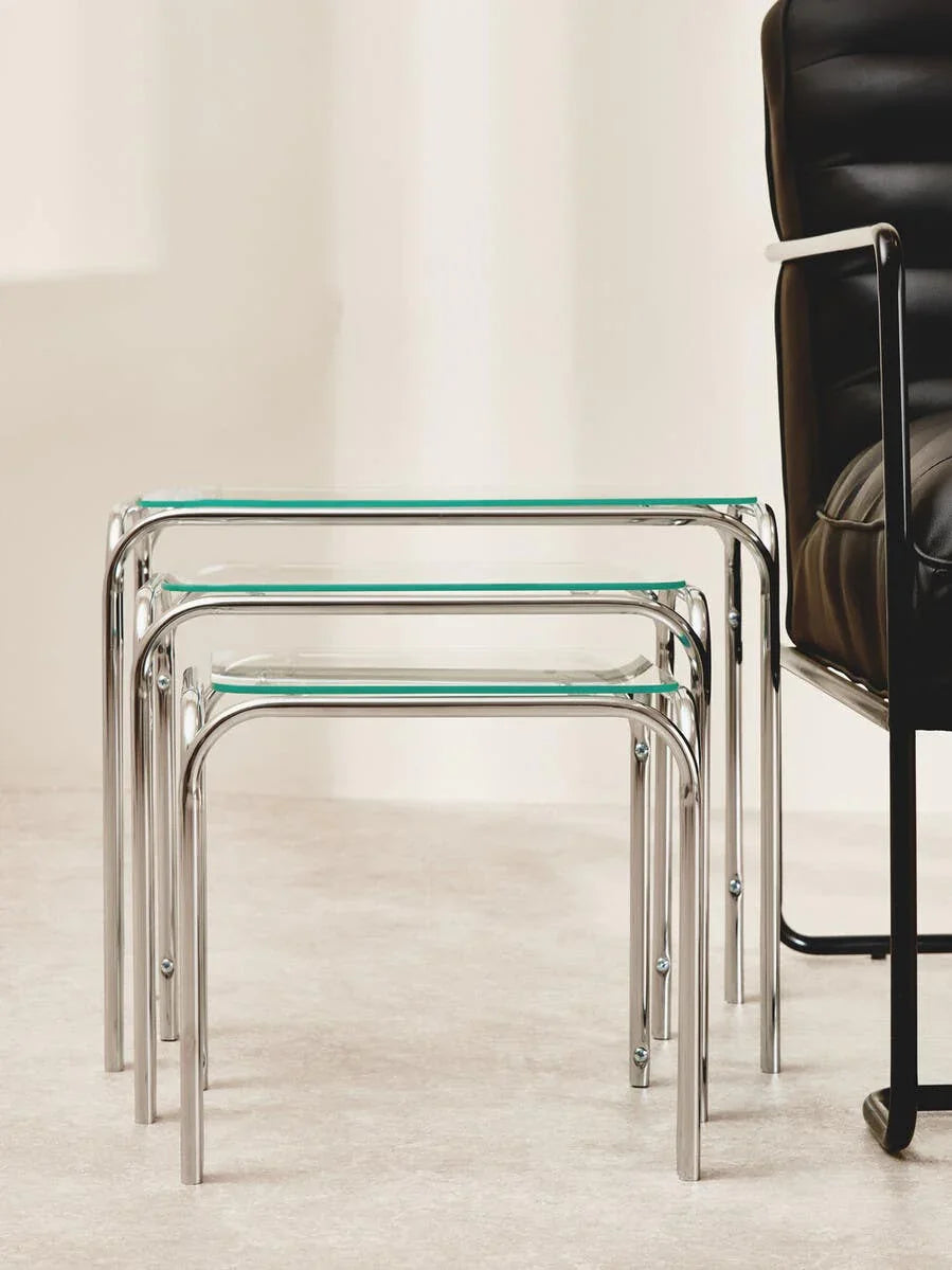 Glass nesting tables with chrome legs beside a black leather armchair on light flooring