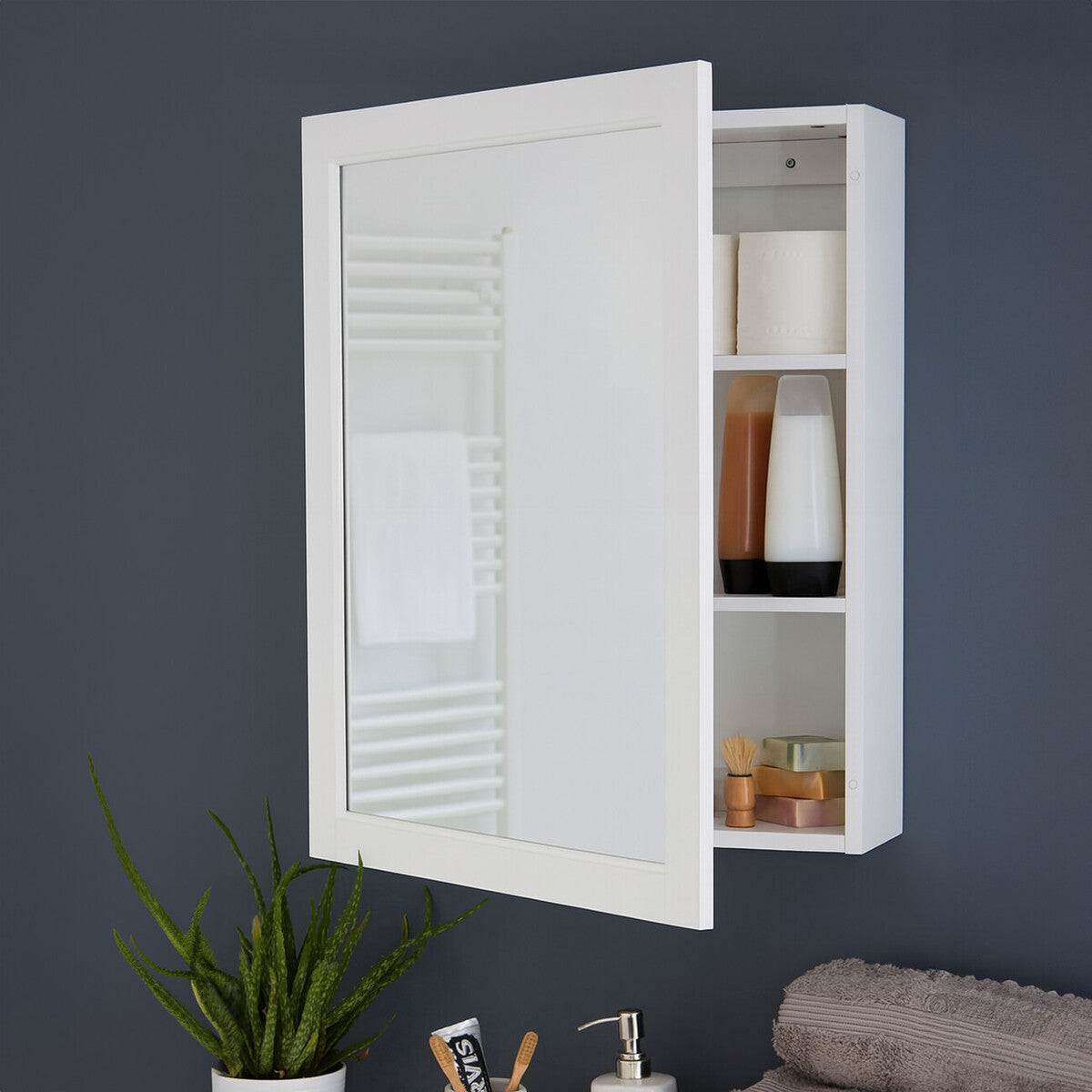 White mirrored wall cabinet with two inner shelves, featuring a louvered door design. The cabinet is mounted on a dark wall, with a small potted plant and a towel visible below it. The shelves display various items, including bottles and containers