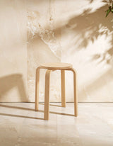A stackable round stool made from tropical Hevea wood, featuring a simple and modern design with clean lines. The stool is positioned on a light-colored surface, casting soft shadows against a neutral backdrop with subtle plant shadows