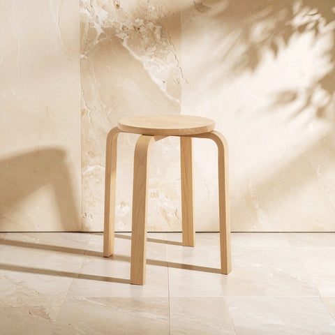 A stackable round stool made from tropical Hevea wood, featuring a light natural finish and smooth, rounded edges. The stool has four sturdy legs and is positioned against a soft, neutral background, casting gentle shadows on the floor