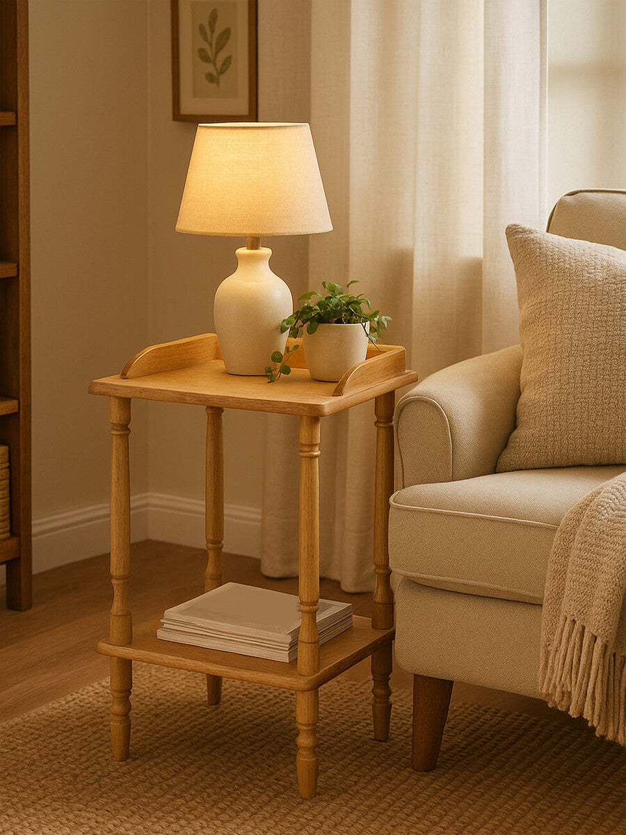 A tropical hevea wood side table with a natural finish, featuring a small lamp on top, a potted plant, and a folded towel on the lower shelf. The table is positioned next to a light-colored sofa in a cozy living room setting with warm lighting