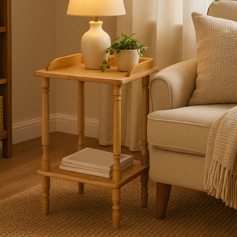 A light-colored Hevea wood side table with a tray-like top, featuring rounded edges and a lower shelf. A small potted plant and a decorative lamp are placed on the table, which stands next to a beige upholstered sofa in a cozy living room setting. Soft natural light filters through sheer curtains, creating a warm atmosphere