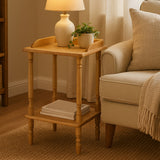 A light-colored Hevea wood side table with a tray-like top, featuring rounded edges and a lower shelf. A small potted plant and a decorative lamp are placed on the table, which stands next to a beige upholstered sofa in a cozy living room setting. Soft natural light filters through sheer curtains, creating a warm atmosphere