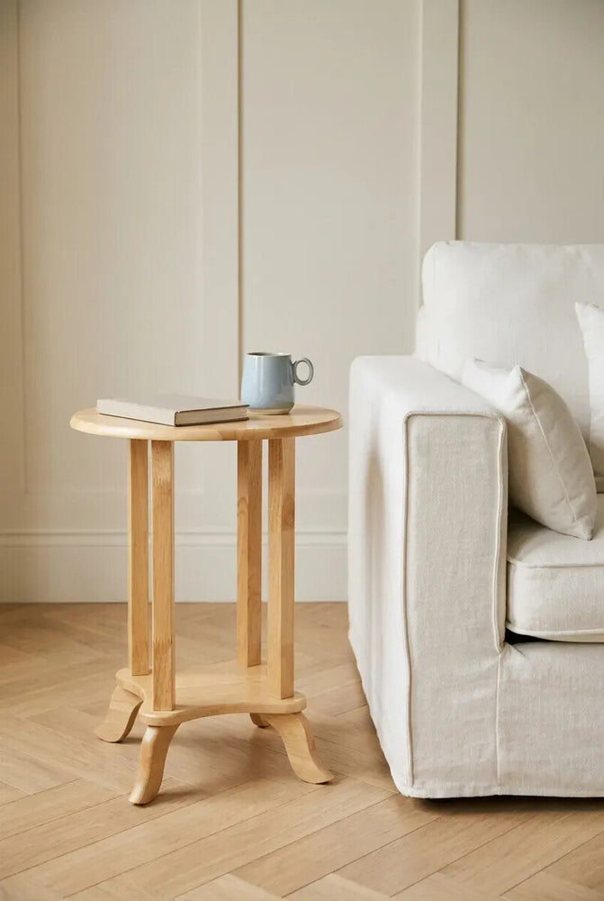 Round side table made from tropical Hevea wood, featuring a light natural finish. The table has a simple, elegant design with a circular top and four sturdy legs, positioned next to a white sofa on a wooden floor. A small blue mug and a book are placed on the table's surface. The background includes a neutral-colored wall with vertical paneling