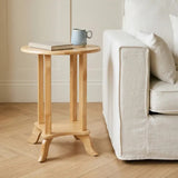 Round side table made of tropical Hevea wood, featuring a light natural finish. The table has a simple, modern design with four sturdy legs and a circular top. A book and a mug are placed on the table, which is positioned next to a light-colored sofa on a wooden floor