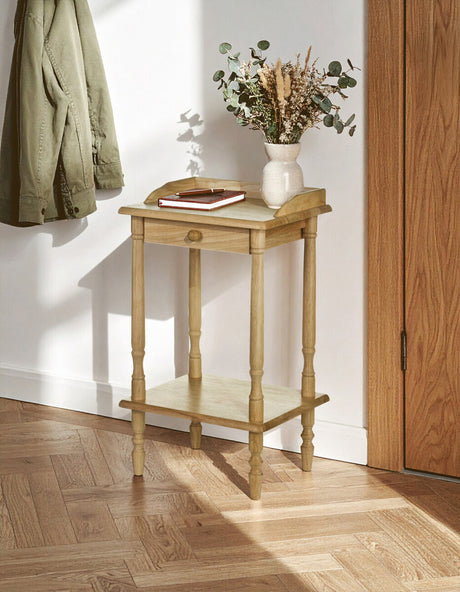 Rectangular side table made from tropical hevea wood, featuring a light wood finish, two levels for storage, and elegantly tapered legs. A small decorative vase with dried flowers and a plate are placed on the top surface, with a wall and wooden door in the background. Soft natural light illuminates the scene, highlighting the table's design and texture