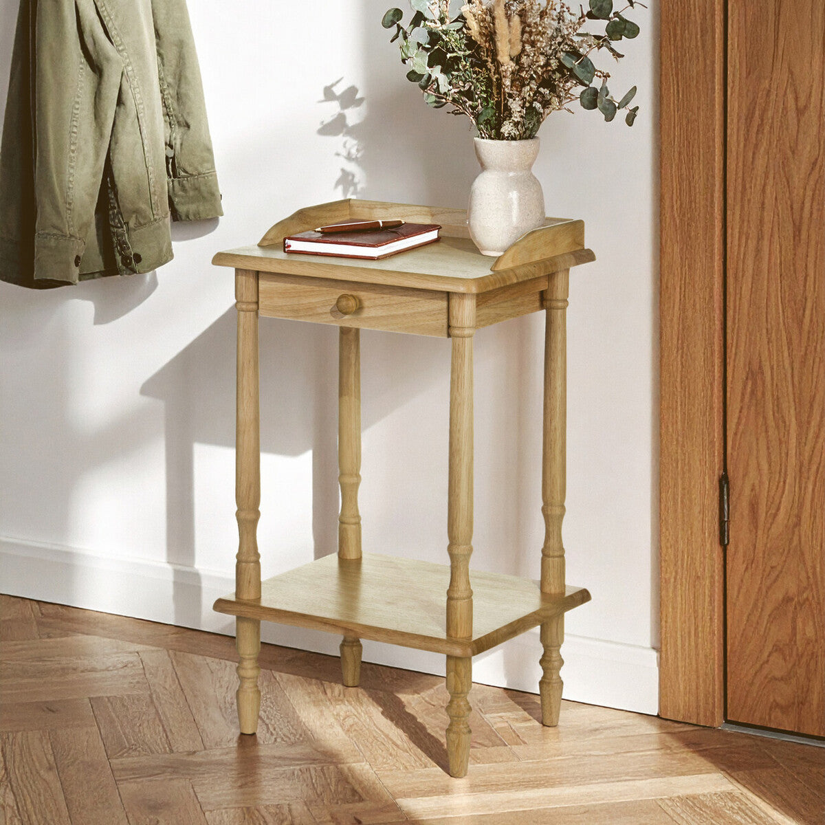 Rectangular side table made from tropical hevea wood, featuring a smooth top surface with a decorative edge, a lower shelf for additional storage, and four slender legs. The table is positioned against a wall with a coat hanging nearby and a vase with dried flowers on top, set on a wooden floor