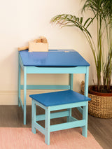 A blue children's desk with a matching stool, both featuring a smooth surface and sturdy legs. The desk has a slanted top and is positioned against a light-colored wall, with a small plant visible in the background. A folded towel or cloth rests on the desk surface