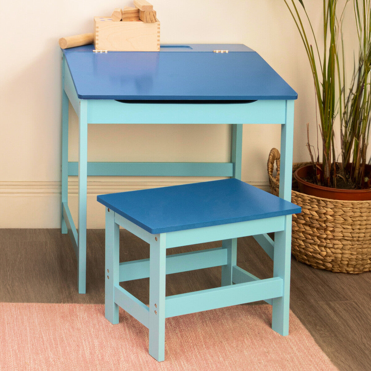 A blue children's desk with a slanted top and a matching stool, both featuring a light blue finish. The desk has a small compartment on top, and the stool is positioned beside it on a light-colored rug, with a potted plant visible in the background