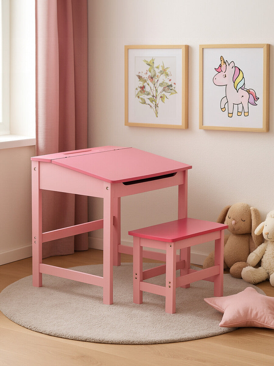 A pink wooden desk with a matching stool, both featuring a simple design, are positioned on a light-colored rug in a cozy room. The desk has a slanted top and a small drawer, while the stool is slightly shorter and has a sturdy frame. Two framed artworks are displayed on the wall, and a plush teddy bear sits nearby, adding a playful touch to the space. Soft natural light filters