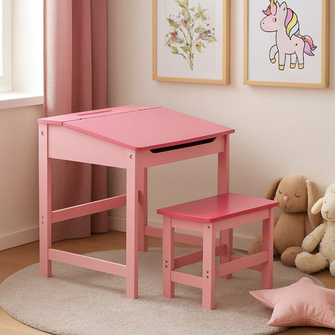A pink children's desk with a slanted top and matching stool, set in a bright room with light-colored walls and a soft rug. The desk features a drawer and is accompanied by plush toys and framed artwork on the walls, creating a playful and inviting atmosphere