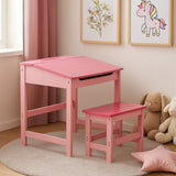 A pink children's desk with a slanted top and matching stool, set in a bright room with light-colored walls and a soft rug. The desk features a drawer and is accompanied by plush toys and framed artwork on the walls, creating a playful and inviting atmosphere